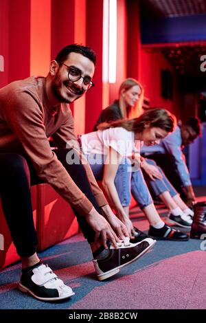 Fröhlicher junger Mixed-Race-Mann in Casualwear und seine Freunde im Hintergrund machen sich bereit, beim Schuhwechsel Bowling zu spielen Stockfoto