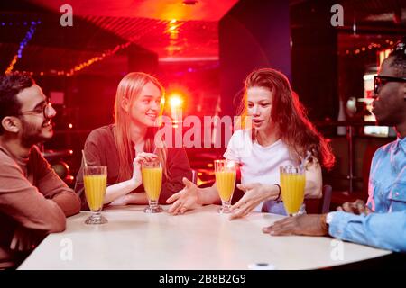 Zwei junge interkulturelle Paare sitzen nach dem Bowling am Tisch im Café im Freizeitzentrum, haben Orangensaft und plaudern Stockfoto