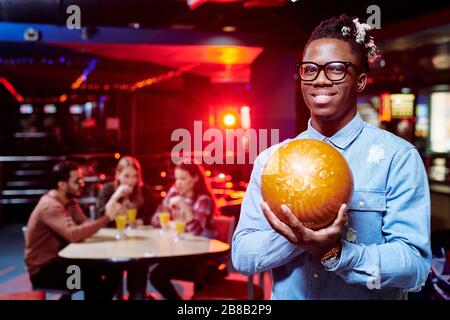 Fröhlicher junger Afrikaner mit Bowling-Ball, der während des Spiels vor der Kamera steht, während seine Freunde Getränke haben Stockfoto