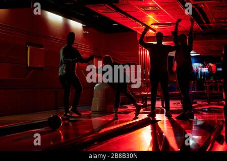 Umrisse von vier aktiven Freunden, die Freude ausdrücken, während einer von ihnen Bowlingball auf eine Gasse wirft, während das Spiel im lesiure Center spielt Stockfoto