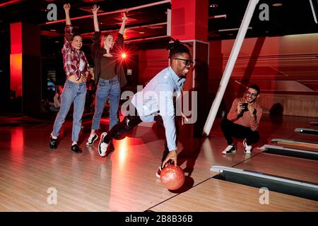 Fröhlicher aktiver Kerl afrikanischer Ethnie, der Bowlingball werfen wird, während zwei glückliche Mädchen Arme hochheben und junger Mann ihn fotografieren Stockfoto