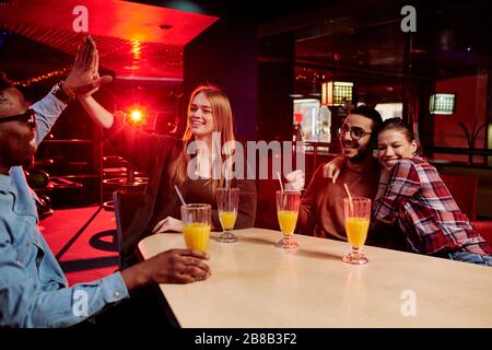 Afrikanischer Kerl und kaukasisches Mädchen, die sich gegenseitig fünf geben, während sie unter Freunden am Tisch sitzen, trinken und den Sieg feiern Stockfoto