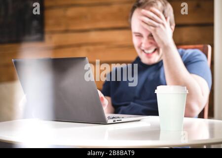 Mann lacht, während er einen Laptop benutzt. Fröhlicher lächelnder Kerl mit Computer im Café, Café oder Zuhause. Lebensfreude, Glück. Lustige Videos ansehen oder streamen. Stockfoto
