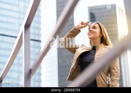 Fröhliche lächelnde Frau in der Großstadt mit Wolkenkratzern. Person, die Sonnenbrille hebt, nach oben schaut und lächelt. Positiver urbaner Lebensstil. Stockfoto