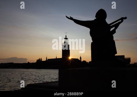 Die Silhouette einer Statue, die die Hand in Richtung Himmel hält und nach etwas bettelt, während die Sonne in einem warmen Sommer hinter dem Stockholmer Rathaus untergeht Stockfoto