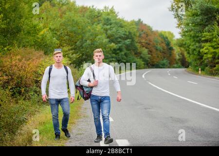 Firmenfreunde Reisende, die auf dem Naturhintergrund der Straße hitchwandern. Fahren Sie in der Nähe des Stadtrandes. Reisende auf dem Weg. Freunde, die hitchhitchhamps unterwegs Beginnen Sie mit dem Wandern ein tolles Abenteuer in Ihrem Leben. Stockfoto