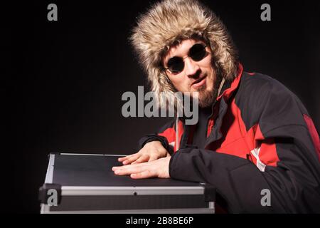 Seitenansicht der gut aussehenden Herren, die auf Runden im Koffer sitzen. Gutaussehender Typ in Sonnenbrille mit Fellhut. Isoliert auf schwarzem Studiohintergrund. Konz Stockfoto
