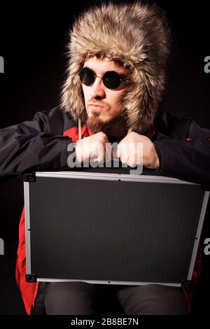 Seitenansicht der gut aussehenden Herren, die auf Runden im Koffer sitzen. Gutaussehender Typ in Sonnenbrille mit Fellhut. Isoliert auf schwarzem Studiohintergrund. Konz Stockfoto