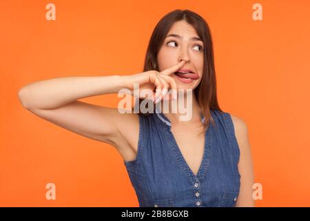 Lustige komische brünette Frau im Denimkleid, die Nase pflückt und Zunge mit humorvollem dummen Ausdruck zeigt, unkultivierte Gewohnheit, schlechtes Umgangskonzept. In Stockfoto