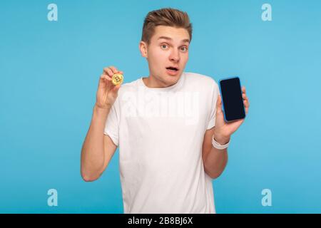 Wow, Mobile Mining App. Porträt des staunenden Mannes im T-Shirt mit goldener Bitcoin und Smartphone in den Händen, überrascht von der Kryptokurrenzie-Technologie. Stockfoto