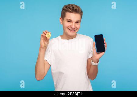 Mobile Brieftasche mit Kryptokurrency. Porträt eines zufriedenen Mannes in legerer weißer T-Shirt mit goldener Bitcoin und Smartphone in den Händen, digitales Bankgeschäft. Stockfoto
