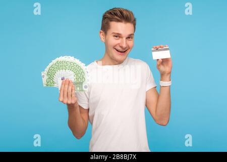 Lustiger fröhlicher lächelnder Mann in weißem T-Shirt mit Kreditkarte und Euro-Banknoten, voller Geldmenge, gerne Bankkredit zum Einkaufen. Stockfoto