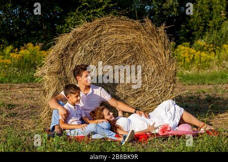 Eine schöne schwangere Frau in einem rosafarbenen Hut hält die Hände auf dem Bauch auf dem Hintergrund eines Heuballens.schwangere Familienfotoaufnahmen in der Natur Stockfoto