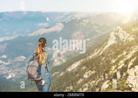 Das junge, schöne Mädchen reist allein im Frühjahr oder Herbst in die Berge, blickt in die Ferne und genießt Natur, Felsen und grüne Wälder, Blick auf die Landschaft, einen Rucksack dahinter und Sportswear, Freiheit und Leichtigkeit Stockfoto