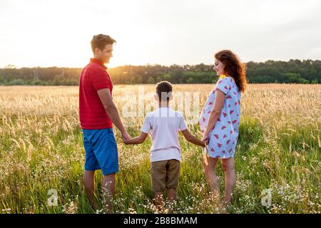 Ein Ehemann, seine Frau und ein junger Sohn posieren bei Sonnenuntergang auf dem Weizenfeld.schwangere Familienfotos schießen in der Natur Stockfoto