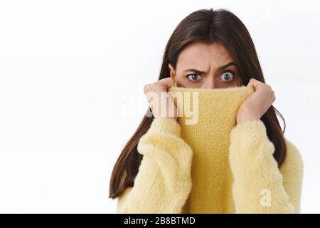 Verdächtige und zweifelhafte brünette Mädchen, die sich mit Pullover im Gesicht verstecken, drücken Unglaube und Zögern mit den Augen aus, sehen unsicher oder freak-out-Gefühl Stockfoto