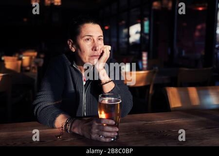 Unglückliche, depressive und einsame Frau mittleren Alters, die mit einem Bierglas an der Bar sitzt. Stockfoto
