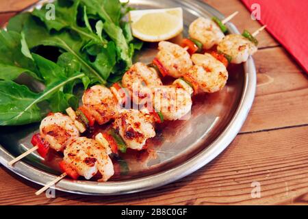 Gegrillte Garnelenspieße auf einem Teller mit Gartenrakete und Zitrone auf einem Holz-Restauranttisch Stockfoto