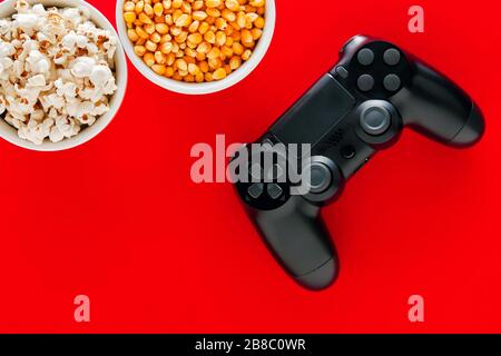 Schüssel Popcorn und eine Schüssel Maiskorn mit einem Gamepad auf rotem Grund. Spielen Sie Konsolenspiele in der Freizeit. Stockfoto