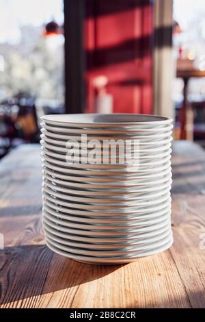 Stapel weißer Teller auf dem Holztisch eines Restaurants oder Cafés unter natürlichem Sonnenlicht am Nachmittag. Stockfoto
