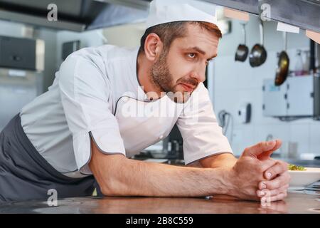 Der konzentrierte kaukasische Koch, der Uniform trägt, ist bereit, seinen Job zu machen. Er wartet auf Aufträge. Seitenansicht. Kochkonzept Stockfoto