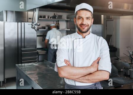 Der junge lächelnde und gutaussehende Koch in weißer Uniform hält seine Hände auf dem Gürtel, der im Inneren der modernen Restaurantküche steht. Vorderansicht. Kochkonzept. Stockfoto