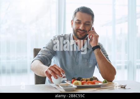 Bärtiger kaukasischer Mann wird Sushi in seinem Büro essen. Er gießt Sojasoße, während er telefoniert. Vorderansicht Stockfoto