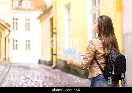 Frau, die in der Stadtstraße spazieren geht und die Karte hält. Rückansicht von weiblichen Touristen und Reisenden mit Rucksack, der während des Urlaubs ein Reiseziel sucht. Stockfoto