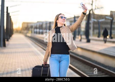 Frau nimmt selfie mit Handy im Bahnsteig am Bahnhof. Lächelnde und fröhliche Dame, die mit Smartphone fotografiert. Stockfoto