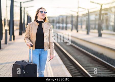 Frau wartet am Bahnhof auf den Zug. Fröhliche lächelnde Dame am Bahnsteig, die auf Bahnfahrt geht. Person mit Pass und Ticket und Blick auf die Spuren. Stockfoto