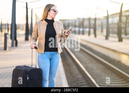 Warten auf Zug. Frau kauft elektronisches Ticket mit Handy auf Plattform. Passagier im Bahnhof mit Gepäck und Koffer. Stockfoto