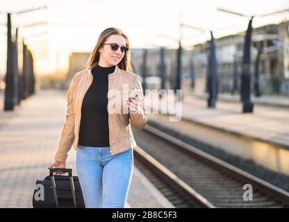 Frau, die im Bahnhof spazieren geht und Smartphone benutzt. Passagier kauft elektronische digitale Tickets mit Handy. Glückliche Dame zieht Koffer- Stockfoto