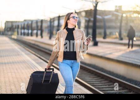 Selbstbewusste Geschäftsfrau zieht Koffer und hält Handy im Bahnhof. Lächelnde Dame, die auf Plattform läuft und Smartphone verwendet. Stockfoto