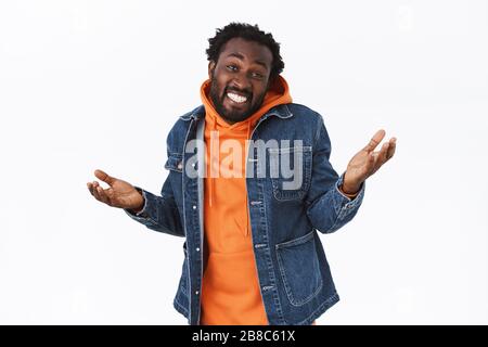 Unbeschwert, entspannt und unbesorgt stiller afroamerikanischer Mann in Denimjacke, orangefarbenem Hoodie, Hände seitlich anhaltenlos oder ahnungslos, lächelnd hochziehen Stockfoto