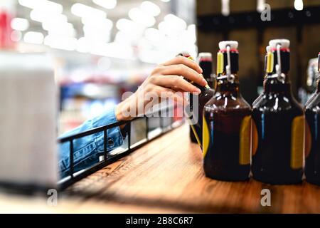 Kunde nimmt Flasche Bier aus dem Regal im Spirituosenladen. Frau, die Alkohol kauft oder Supermarktmitarbeiter, die Regale in einem Getränkegang befüllen und befüllen. Stockfoto