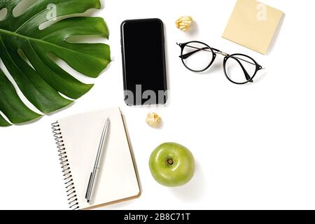 Schreibtisch mit Blick auf den Bürotisch. Arbeitsbereich mit Stift, Monstera-Blättern, Brillen, Notizbuch, apple, modernes Smartphone mit Kopierbereich auf weißem Hintergrund mi Stockfoto