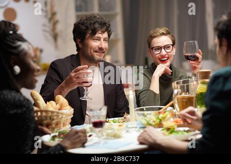 Junges lächelndes Paar, das Wein am Tisch trinkt und die Gesellschaft ihrer Freunde beim Feiertagsabendessen zu Hause genießt Stockfoto