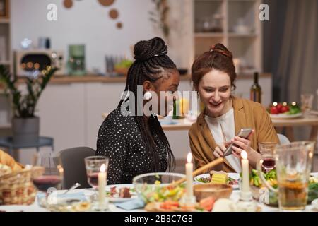 Zwei junge Frauen, die Fotos auf dem Handy ansehen und lächeln, während sie zu Hause am Feiertagstisch sitzen Stockfoto