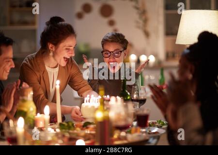 Fröhliche junge Frau in Brillen ist so begeistert von Geburtstagskuchen mit Kerzen, während sie zusammen mit ihren Freunden am Tisch sitzt Stockfoto