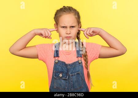 Möchten Sie nicht zuhören. Portrait des kapriziösen kleinen Mädchens in Denim-Overalls, die mit geschlossenen Ohren stehen, um den Rat der Eltern nicht zu hören, und die Erziehung ignorieren. Stockfoto