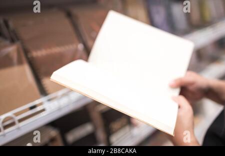 Leeres weißes Blatt Notizbuchpapier ohne Zeile zum Schreiben von Textnotizen. Die Hand einer Frau hielt ein Buch mit leerem Papier auf einem braunen Korkbezug. Stockfoto