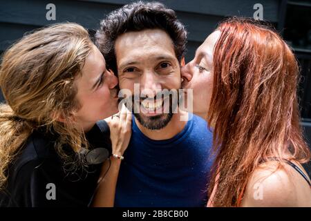 Zwei junge Frauen, die einem jungen Mann unterschiedlicher Ethnie mit Bart und Schnurrbart auf der Straße einen Kuss auf jede Wange geben Stockfoto