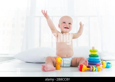 Charmantes fröhliches Kind in einer Windel lacht und spielt, während es in seinem gemütlichen Zimmer sitzt. Das Konzept einer gesunden, glücklichen Baby- und Kinderbetreuung Stockfoto