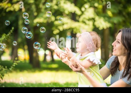Lächelnde junge Mutter, die ihrer kleinen Tochter hilft, am Sommerfreitag Blasen im Park zu fangen. Fröhliches Familienzeit-Konzept Stockfoto