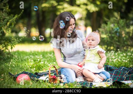Lächelnde junge Mutter, die ihrer kleinen Tochter hilft, am Sommerfreitag Blasen im Park zu fangen. Fröhliches Familienzeit-Konzept Stockfoto