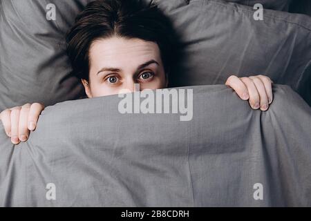 Draufsicht auf fröhliche junge Frau versteckt sich unter weicher grauer Decke, hat Spaß im Bett, schaut auf die Kamera, freut sich guten Morgen, ruht im Bett. Ansicht von oben. Stockfoto