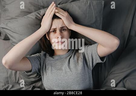 Unglückliche junge brünette Frau im Bett liegend, Kamera ansehen, Tempel nah berühren, müde Frauen leiden an Kopfschmerzen oder Migräne, fühlen sich unwohl Stockfoto