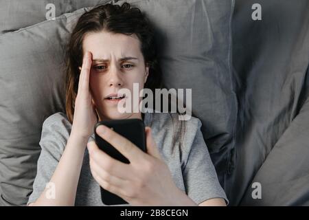Draufsicht über unglückliche Frauen, die auf Kopfkissen im Bett liegen und auf dem Smartphone-Bildschirm schlechte Nachrichten lesen, überschlafen, junge Frauen mit offenem Mund verärgern, fassungslos Stockfoto