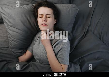 Draufsicht auf ungesunde kranke junge Frau, die auf dem Bett liegt, an Halsschmerzen leidet, den Hals berührt, schmerzhaftes Gefühl hat Stockfoto