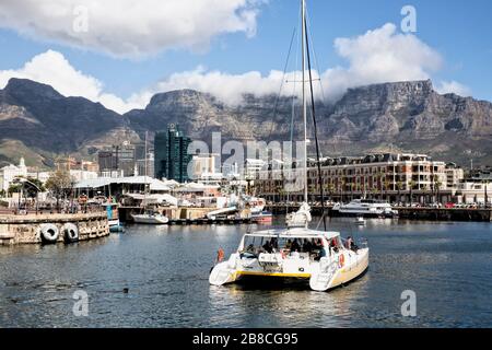 Ein mit Touristen beladener Katamaran fährt in Richtung Cape Grace Hotel an der V & A Waterfront, während der Tafelberg von hinten auf die Stadt blickt Stockfoto